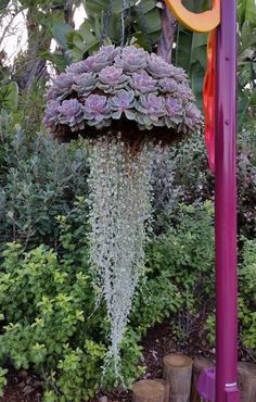 an umbrella made out of flowers in the middle of some bushes and trees with purple ribbons hanging from it