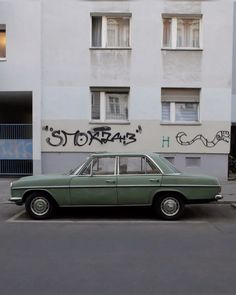 an old green car parked in front of a building with graffiti on it's side