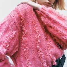 a woman wearing a pink sweater with ruffles on the sleeves and shoulders is posing in front of a white wall