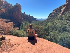 a woman sitting on top of a red rock next to a lush green forest covered hillside