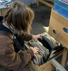 a woman is looking through an open cardboard box filled with magazines and records on the floor