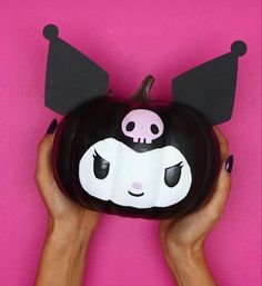 a hand holding a black and white pumpkin with ears on it's head, against a pink background