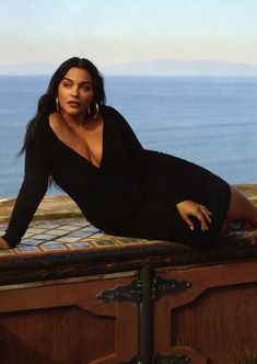 a woman laying on top of a wooden trunk next to the ocean with her legs crossed