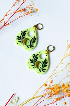 two green earrings sitting on top of a white table next to dried flowers and twigs