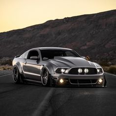 the front end of a silver mustang on a road with mountains in the back ground