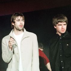 two young men standing next to each other in front of a dark background, one holding a cell phone