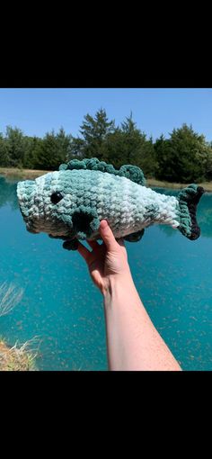 a hand holding up a fake fish in front of a body of water with trees in the background