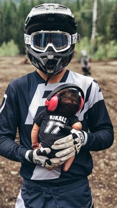 a man holding a baby wearing a helmet and goggles on his head while standing in the dirt