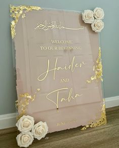 a wedding welcome sign with flowers on the bottom and gold foiling around it, in front of a blue wall