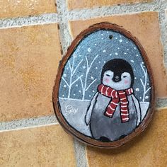 a painted rock with a penguin wearing a scarf on it's head and snowing trees in the background