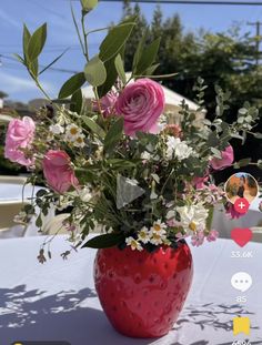 a red vase filled with lots of flowers on top of a white cloth covered table