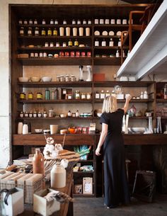 a woman standing in front of a wooden shelf filled with lots of bottles and jars