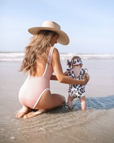 a woman in a bathing suit and hat sitting on the beach next to a baby