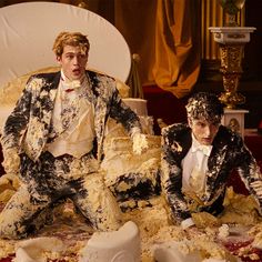 two men sitting on top of a bed covered in fake food and plastic wrappers