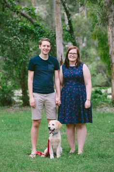 a man and woman standing next to a dog in the grass with trees behind them