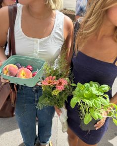 two women standing next to each other holding flowers and fruit