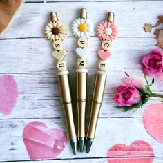 three pens sitting on top of a wooden table next to pink flowers and hearts shaped erasers