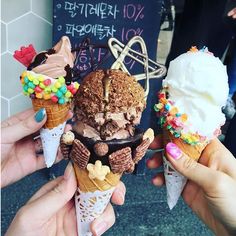 three people holding ice cream cones with toppings on them in front of a sign