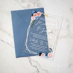 two blue and white wedding cards with floral designs on them, sitting on a marble surface