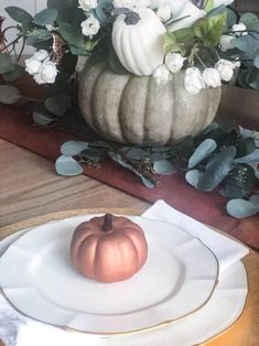 a white plate topped with a pumpkin next to a vase filled with flowers and greenery