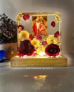 a glass frame with flowers and a couple in the center on a wooden stand next to some plants