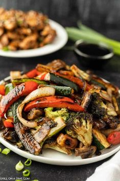 a plate full of stir fried vegetables with sauce on the side and another plate in the background