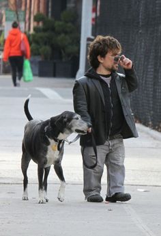 a man talking on a cell phone while walking his dog down the street in front of him