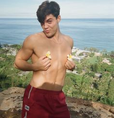 a man with no shirt on standing next to the ocean and holding flowers in his hand