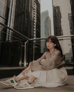 a woman sitting on the ground in front of tall buildings with her legs crossed and looking off into the distance