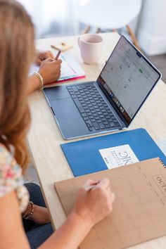 a woman sitting at a table with a laptop and notebook in front of her, writing on a piece of paper