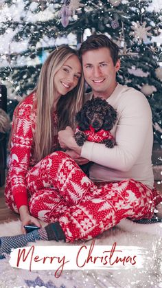 a man and woman sitting in front of a christmas tree with a dog on their lap