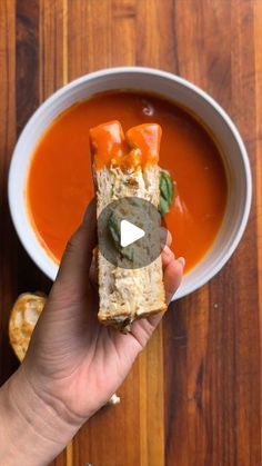 a person holding a sandwich in front of a bowl of tomato soup on a wooden table