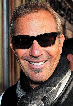 a man wearing sunglasses and a scarf smiling for the camera with another man in the background