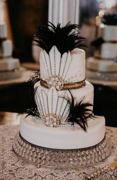 a three tiered white cake with black feathers and beads on the top, sitting on a table