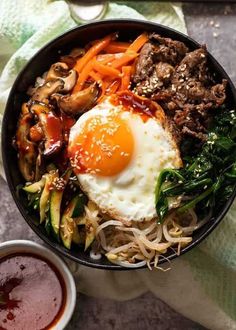 a bowl filled with meat, vegetables and an egg on top of it next to dipping sauce