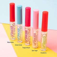 four different lip glosses are lined up on a yellow and pink surface, with the names of each product