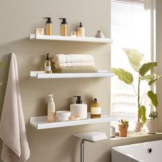 two white shelves above a bath tub filled with personal care items and plants in front of a window