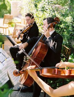 two men in suits playing violin and another man wearing a tuxedo sitting on a bench