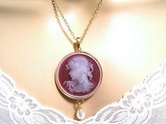 "Beautifully detailed round Victorian cameo necklace with a pendant of a neoclassical woman and flowers against a cherry red background. Hanging below like a drop of rain is a cream Swarovski pearl. The cameo is set in a round shiny gold plated pendant. Length: 17 inches, Locket Pendant: 3.15 inches Arrives with a Marty White card in a lovely decorative bag and bubble wrap packaging. Facebook: http://www.facebook.com/pages/Marty-whitedesigns/159425327440004 ** Please \"Favorite\" my shop and com Red Medallion Necklace For Formal Occasions, Cameo Round Pendant Necklace For Wedding, Wedding Necklaces With Cameo Round Pendant, Red Cameo Jewelry For Wedding, Red Cameo Necklace For Gift, Red Cameo Jewelry Gift, Cherry Red Background, Woman And Flowers, Victorian Cameo