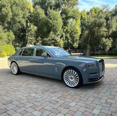 a grey rolls royce parked in front of some trees and bushes on a brick driveway