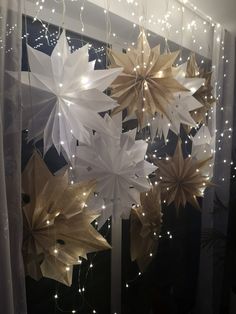 some white and gold paper stars hanging from the ceiling in front of a window with lights