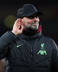 an older man with a beard wearing a black liverpool united jacket and hat, holding his hand to his ear