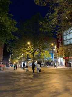 people walking down the street at night in an open area with tall buildings and trees