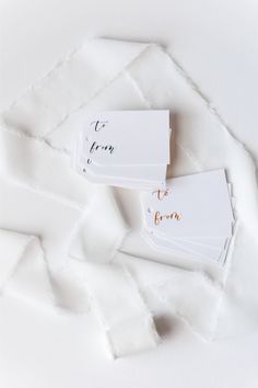 two place cards with writing on them sitting next to each other in white fabric and gold foil