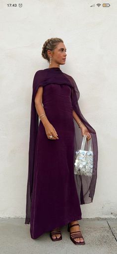 a woman wearing a purple dress and holding a white purse standing in front of a wall