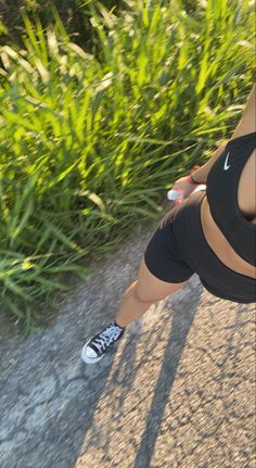 a woman riding a skateboard down a street next to tall grass and bushes on either side of her