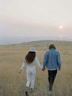 two people holding hands while walking through tall grass with the sun setting in the background