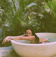 a woman in a bathtub surrounded by greenery and palm trees, posing for the camera