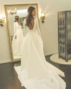 a woman standing in front of a mirror wearing a white wedding dress with long sleeves