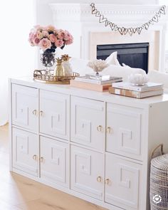 a living room with white furniture and pink flowers on top of the dresser in front of the fireplace